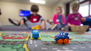 Billede med to legetøjsbiler i forgrunden på et biltæppe, og børnehavebørn ude af fokus i baggrunden